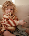 A young child with curly blonde hair, dressed in brown and beige stripes, sits on a cream couch holding a gray moon toy and blanket, using a FRIGG Rope Silicone Pacifier—one of the baby essentials from FRIGG’s 2-pack collection.