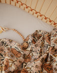 A mushie Organic Cotton Muslin Swaddle Blanket with a floral pattern and a beaded pacifier clip are placed on a white sheet inside a light wooden crib.