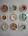 Nine sets of mushie Square Dinnerware Plates, made from BPA-free silicone in neutral and pastel tones, are arranged in a grid on a light surface. Each children’s set includes a plate or bowl with a matching fork or spoon.