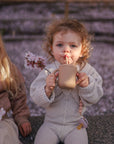 Two young children sit outdoors among blossoms. One, with curly hair, sips from a cup and holds flowers, her mushie Silicone Pacifier Clip | Hera attached nearby. The other child, wearing glasses, is partially visible beside her.
