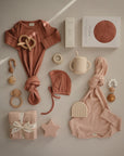 A flat lay of baby items in soft pink and beige, featuring a mushie Ribbed Baby Bonnet, knotted onesie, bibs, teething toys, sippy cup, rainbow and star-shaped toys, pacifier clip, boxed goods, and folded cloth.