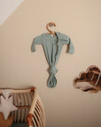 A sage green knotted baby gown hangs on a wooden hanger above a rattan crib. Nearby, a cloud-shaped mirror, star pillow, and the mushie Ribbed Baby Bonnet in organic cotton complete the cozy nursery scene.