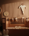 A cozy nursery with a wooden crib, neutral bedding, and soft toys. A mushie Ribbed Baby Bonnet hangs beside baby clothes on the wall, while shelves display decor and sunlight brightens the inviting space.