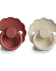 The FRIGG Daisy Silicone Pacifier 2-Pack by FRIGG features two medical grade silicone pacifiers—one muted red, one light cream—with a safe, comfortable scalloped-edge design, shown side by side on a white background.