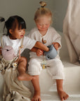 Two young children sit together on white steps, smiling and playing, with a soft blanket and FRIGG Daisy Night Natural Rubber Pacifier 2-Pack glowing in the dark—natural light from the side adds to the cozy scene.