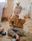 A young child with blonde hair in a bun sits on a woven rug, playing with wooden blocks beside the mushie Silicone Pacifier Clip | Eva—made from food-grade silicone—combining playtime with practical pacifier accessories.