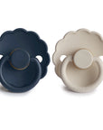 The FRIGG Daisy Natural Rubber Pacifier 2-Pack features two daisy-shaped pacifiers with scalloped edges, one dark blue and one cream, each with a round handle, circular shield with two small holes, and a natural rubber latex nipple.