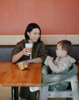A woman holding a coffee cup smiles at a toddler in a high chair at a wooden table. The toddler reaches for their Mushie Trainer Sippy Cup with easy-grip handles, sitting by a container of fruit against a red and tan wall.