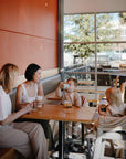 Two women sit at a café table with two young children—one drinks from the Mushie Trainer Sippy Cup, featuring easy-grip handles and non-toxic materials, while the other sits in a high chair as sunlight fills the bright, cheerful room.