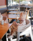 Two toddlers sit at wooden tables in a sunlit café. One, in a high chair, holds a Mushie Trainer Sippy Cup with easy-grip handles while the other reaches for food, their neutral outfits enhancing the calm atmosphere.
