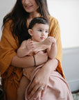 A woman in a mustard robe holds a baby dressed in pink, who sits on a Mushie Ribbed Baby Blanket and holds a beige toy. The softly lit background is out of focus.