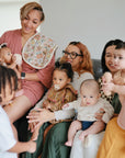 Three women sit on a couch, each holding a baby wrapped in a Mushie Ribbed Baby Blanket. A young child stands beside them. The group shares smiles and warm moments together in a cozy, sunlit room.