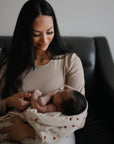 A woman with long dark hair, in a beige top, sits on a dark couch gently cradling her newborn wrapped in the mushie Organic Cotton Muslin Swaddle Blanket, smiling tenderly down at the baby.