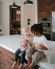 Two young children sit on a modern kitchen counter, one holding a Mushie Trainer Sippy Cup with easy-grip handles and non-toxic materials, and the other holding a toy teapot as they play or talk together.