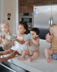 Four toddlers sit side by side on a kitchen counter, each holding a Mushie Trainer Sippy Cup with handles. Dressed in neutral outfits, they look in various directions—one reaches out and another sucks on a pacifier.