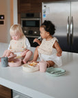 Two toddlers sit on a kitchen counter, happily snacking and drinking from Mushie Trainer Sippy Cups with easy-grip handles. Both cups are non-toxic. A refrigerator and other kitchen appliances appear in the background.