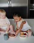 Two toddlers sit on a kitchen counter with pastel plates and cups. One drinks from the Mushie Trainer Sippy Cup, featuring easy-grip handles, as they share food and play with utensils, enjoying their non-toxic sippy cup.