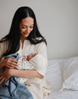 A woman with long dark hair sits on a bed, smiling down at her newborn, who is wrapped in a mushie Organic Cotton Muslin Swaddle Blanket and wearing a hat. The serene scene features white bedding and a neutral background.