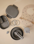 A flat lay of gray and beige baby feeding items, including a divided lunchbox, buttoned bib, teething rings, pacifier, two bowls with utensils, and a Mushie Trainer Sippy Cup with easy-grip handles, all on a light surface.