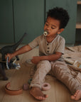 A young child with a FRIGG Daisy Night Silicone Pacifier from FRIGG sits on the floor playing with toy dinosaurs and stacking rings, surrounded by soft brown and beige tones.