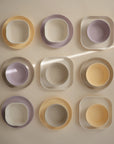 Nine mushie Square Dinnerware Plates in pastel yellow, lavender, and white are arranged in a grid on a beige background. A diagonal beam of sunlight highlights this BPA-free set featuring both round and square shapes.