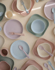 Mushie Round Dinnerware Bowls, BPA-free and in soft pastel colors like pink, blue, green, and beige, are neatly arranged on a light surface with matching cups, spoons, and forks. Most pieces are round or square.
