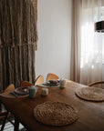 A wooden dining table is set with round woven placemats, ceramic dishes, a teapot, and mugs. The Mushie Trainer Sippy Cup with easy-grip handles sits nearby. Sunlight filters through sheer curtains; chunky textile art decorates the wall.