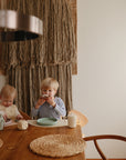 Two children sit at a wooden dining table; one eats a sprinkled donut, the other draws. The table has cups, plates, woven placemats, and a Mushie Trainer Sippy Cup with easy-grip handles. Yarn wall art decorates the background.
