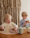 Two young children sit at a table eating. The toddler in front wears a bib, using a fork with the mushie Silicone Suction Plate, while the older child in the background holds toast. Cups and a toy block are also on the table.