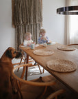Two young children eat at a wooden dining table, sipping from Mushie Trainer Sippy Cups with easy-grip handles, while a large brown dog sits nearby. Sunlight highlights the woven wall hanging behind them.