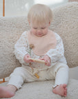 A blonde-haired baby sits on a textured chair, playing with a wooden toy. Nearby rests the mushie Silicone Pacifier Clip | Eva, which matches the baby's light pink bib and white polka-dotted clothes.