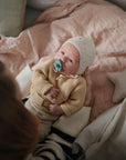 A baby in a beige knitted outfit and bonnet lies on a soft bed, using the FRIGG Daisy Silicone Pacifier from FRIGG, while an adult cradles them and a star-shaped pillow rests nearby.