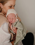 A woman cradles her sleeping newborn, bundled in a light green blanket and beige bonnet. On her shoulder rests a mushie Organic Cotton Muslin Burp Cloth from the 2-Pack, enhancing the warmth and tenderness of the moment.