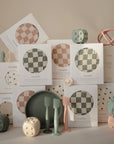 A display of baby products, including mushie’s boxed muslin swaddles in checkered green and pink, silicone utensils, plates, stacking toys, pastel dice, teething rings, and an extra soft muslin changing pad cover.