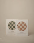 Two boxes of mushie Extra Soft Muslin Changing Pad Covers—one green and white checkered, one pink and white checkered—are shown against a neutral beige background.