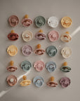 A neatly arranged grid of 20 Mushie FRIGG Rope Natural Rubber Pacifier 2-Packs in pastel and muted colors, each with a natural rubber latex nipple, displayed on a light surface with soft shadows—perfect for baby essentials.
