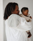 A woman in a white shirt holds a baby with a pacifier, facing right in soft light. On her shoulder is the mushie Organic Cotton Muslin Burp Cloth (2-Pack); the baby wears a light ribbed outfit.