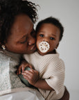 A woman lovingly kisses a baby in a beige knit outfit, holding a pacifier. Nearby are mushie Organic Cotton Muslin Burp Cloths (2-Pack), offering gentle comfort against the plain background.