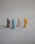 Four mushie Finger Toothbrushes in pastel purple, blue, white, and yellow stand upright on a light surface. These gentle silicone brushes are perfect for cleaning baby teeth against a soft, neutral background.