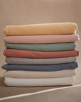 A neatly stacked pile of mushie Baby Wraps in solid colors like yellow, pink, green, gray, red, blue, beige, and white—crafted from organic cotton—set against a neutral brown background. Perfect for a soft and breathable baby wrap.