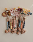Five FRIGG Daisy Natural Rubber Pacifier 2-Packs with colorful silicone and wooden beaded clips are arranged in a row, featuring natural rubber latex nipples and beads in pastel and muted tones against a neutral background.