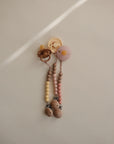 Two mushie Silicone Pacifier Clip | Eva accessories hang against a neutral wall: one beige with yellow beads, the other pink with peach beads. Both feature wooden accents on the clip ends, softly lit in the image.