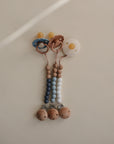 The Mushie FRIGG Rope Natural Rubber Pacifier 2-Pack features pacifiers with natural rubber latex nipples and beaded silicone and wooden clips in blue, gray, and white, displayed neatly against a beige background.