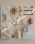 A neatly arranged flat lay of neutral-toned baby items—including bottles, muslin cloths, skin care products, a bonnet, food-grade silicone teething rings, a pacifier case, and the mushie Silicone Pacifier Clip | Eva—is displayed on a light background.