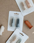Mushie Finger Toothbrushes in neutral colors are displayed on a beige surface, both packaged and loose. Each soft silicone teething brush fits over your finger for gentle cleaning of baby's teeth.