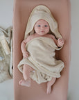 A baby lies on the mushie Extra Soft Muslin Changing Pad Cover in pink, wrapped in a cozy beige hooded towel. Nearby are a striped cloth, cream tube, bottle, and pacifier clip as the baby gazes up with wide eyes.
