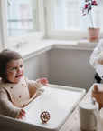A smiling baby in a knit bonnet sits in a high chair, reaching for the Mushie Trainer Sippy Cup with easy-grip handles. To the right, an adult holds a small dog as sunlight streams through the window.