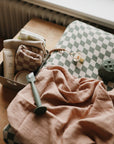 A wooden table holds a mushie Extra Soft Muslin Changing Pad Cover, a peach muslin cloth, a pacifier clip, a green teether, and a small box with baby essentials like lotion, a washcloth, and a pouch.