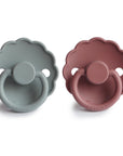 The FRIGG Daisy Silicone Pacifier 2-Pack includes two daisy-shaped, scallop-edged pacifiers—one muted blue, one muted mauve—with round handles and FRIGG branding. Made of medical-grade silicone for safety and comfort.
