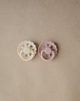 Two FRIGG Andersen Fairytale Silicone Pacifiers, one cream and one light pink, with decorative edges and heart-shaped cutouts, are placed side by side on a plain brown surface.